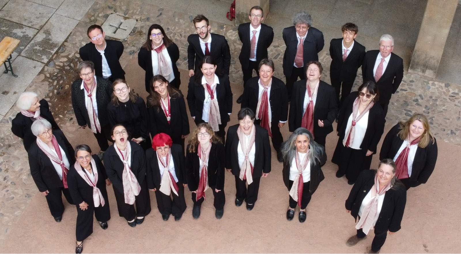 Les choristes du Chœur Polyphonique du Forez, vus de dessus dans une cour pavée, en tenue noire et écharpes roses, le visage levé vers l’objectif